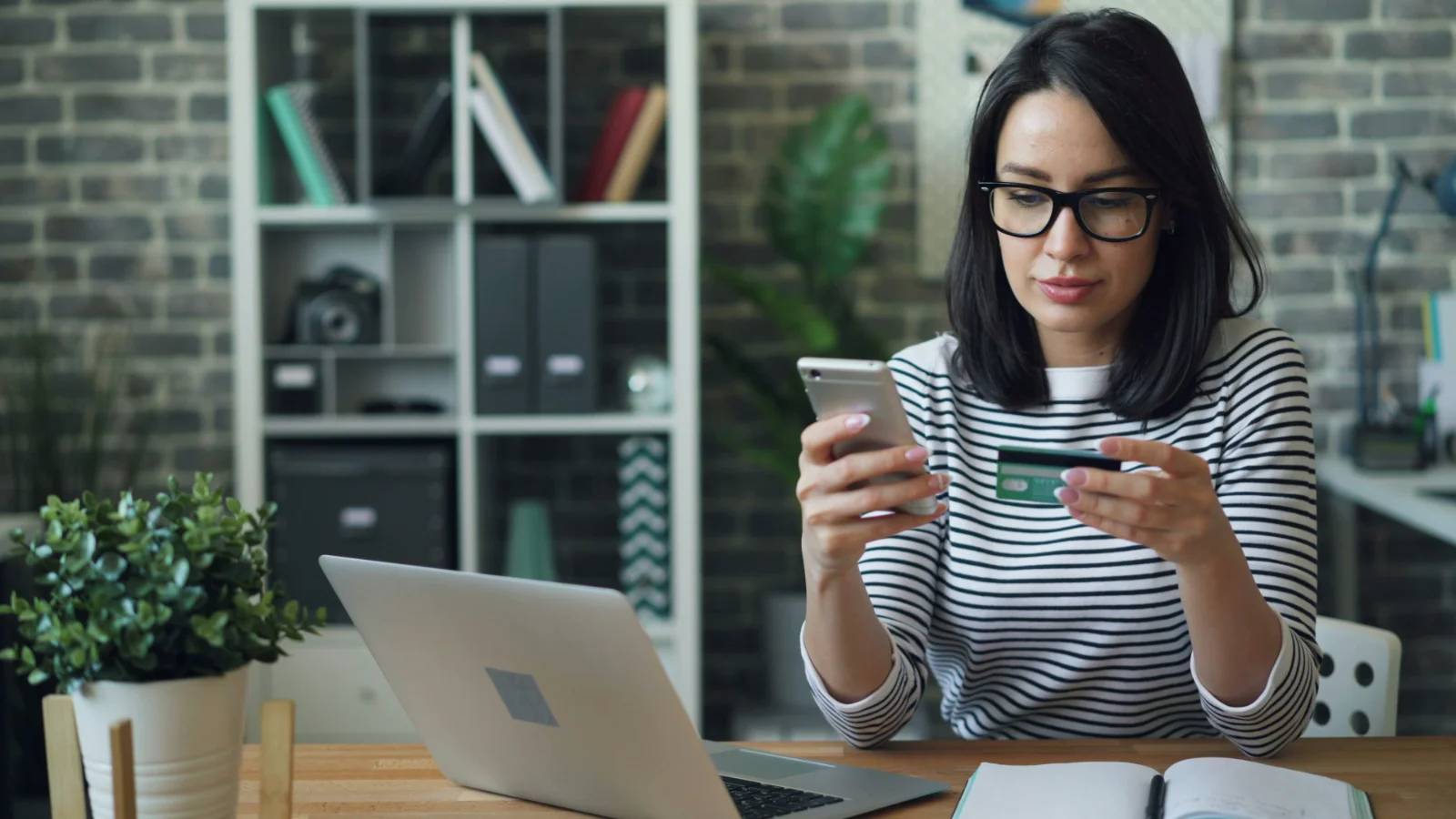 A woman sitting at a desk holding a phone and a credit card, entering payment details with a laptop open in front of her.