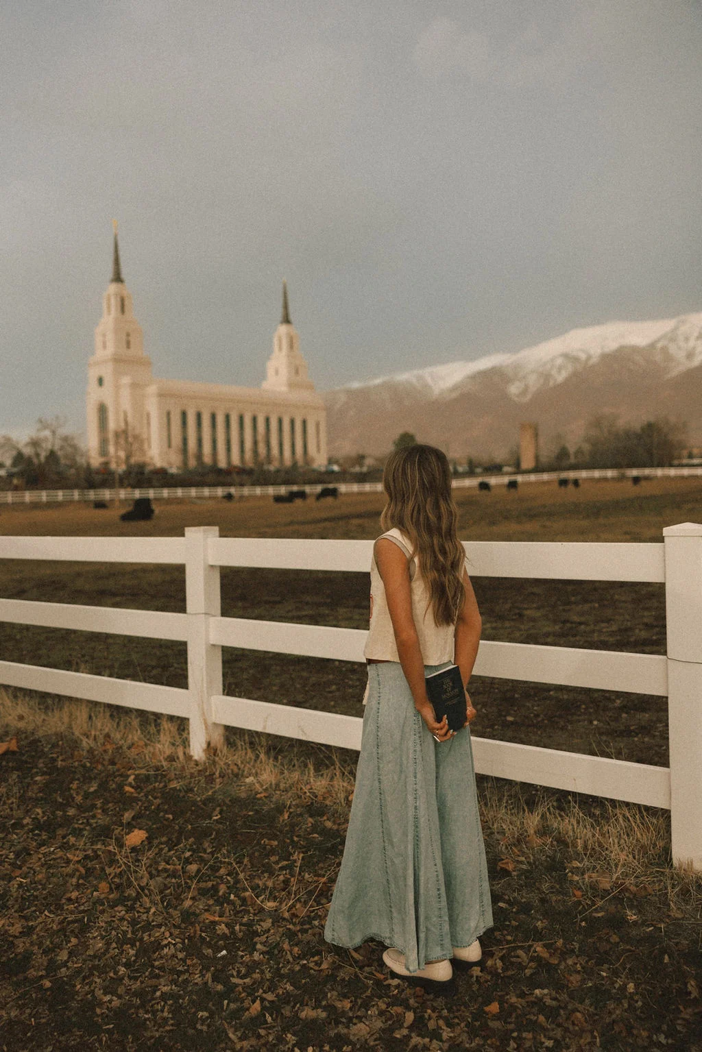 A young woman seen from behind, holding a book, standing at a white fence overlooking a field with a large temple building and mountains in the background.
