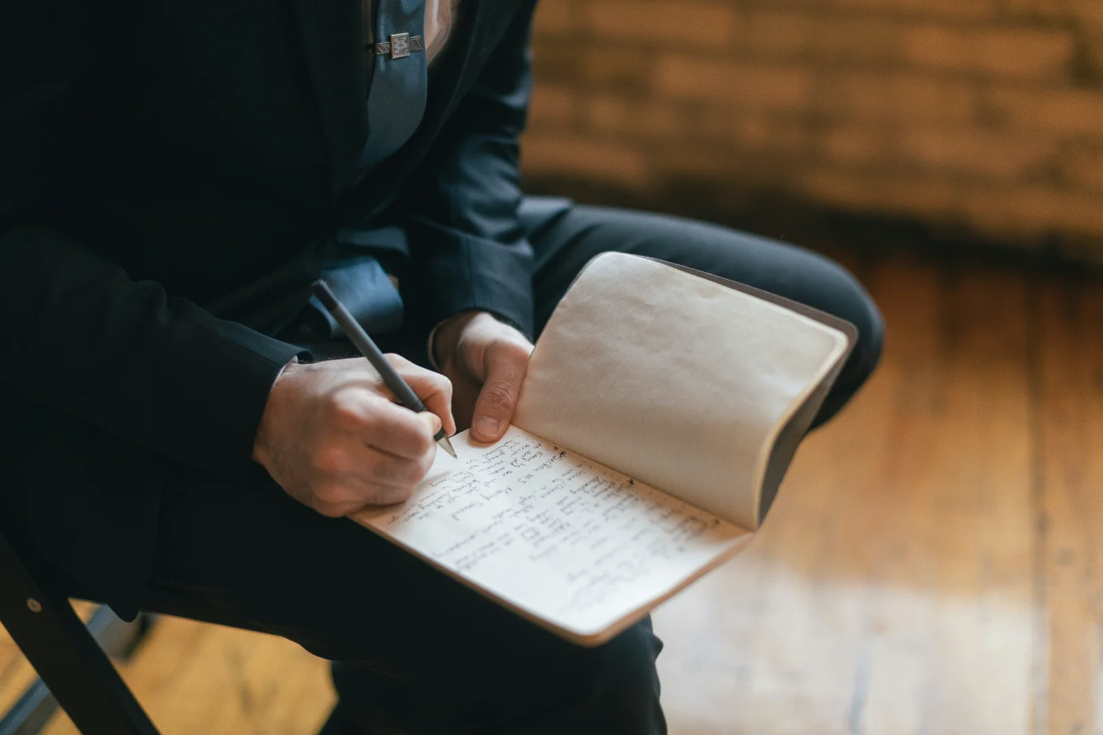 Close-up of a man in a suit writing in a small notebook while seated, with a wooden floor visible in the background.