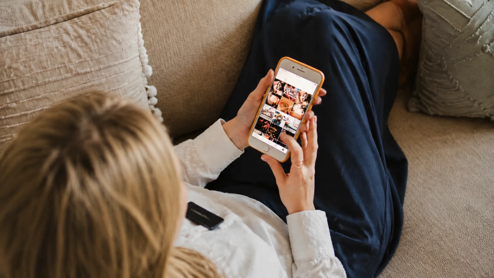 Overhead view of a person scrolling through a photo grid on a smartphone while seated on a couch.
