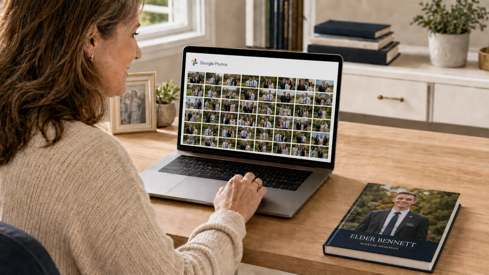 A woman looking at a grid of photos open in Google Photos on a laptop, next to a printed missionary keepsake book on a wooden desk.