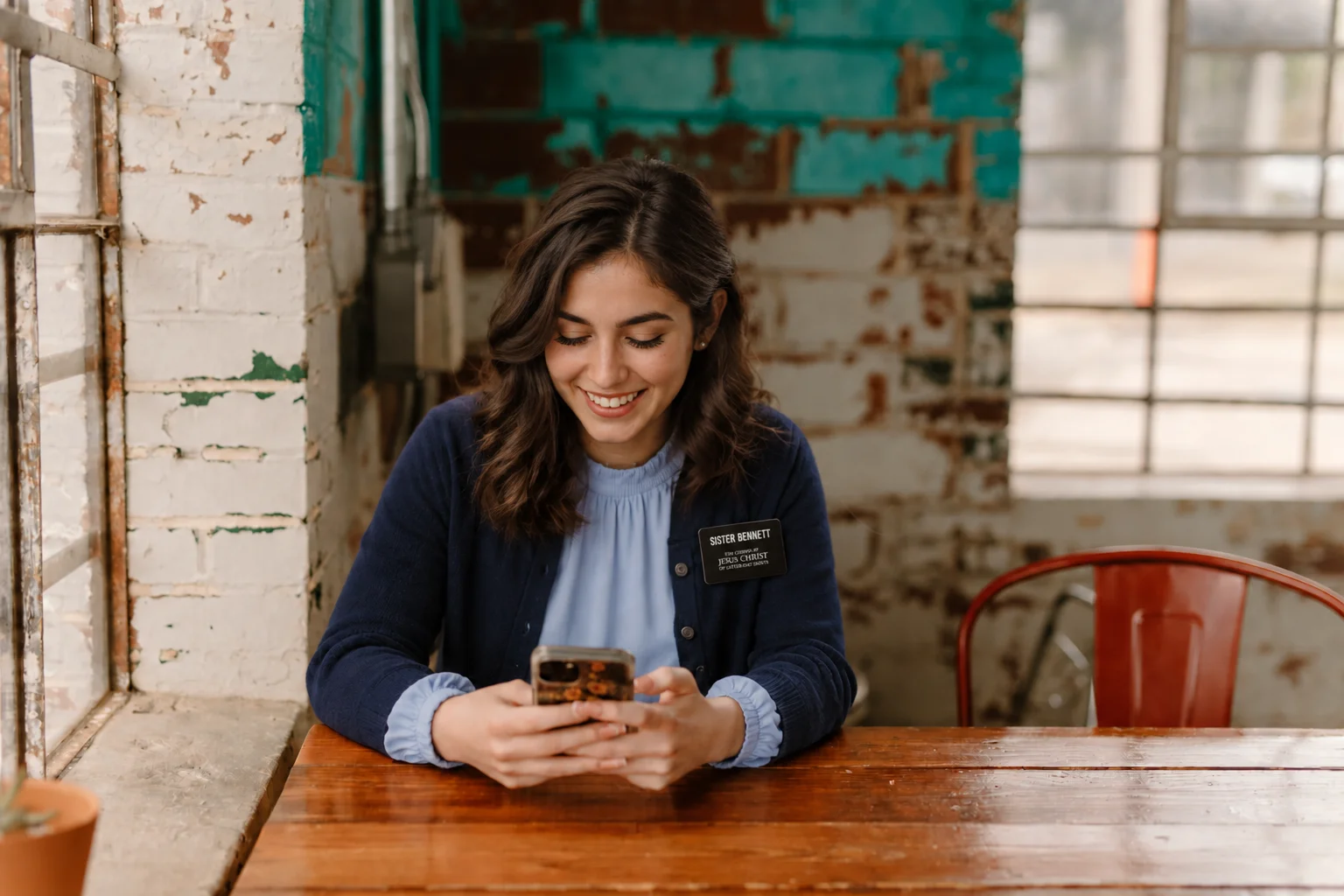 Sister missionary wearing a blue cardigan and name badge smiles while using her phone at a café table.