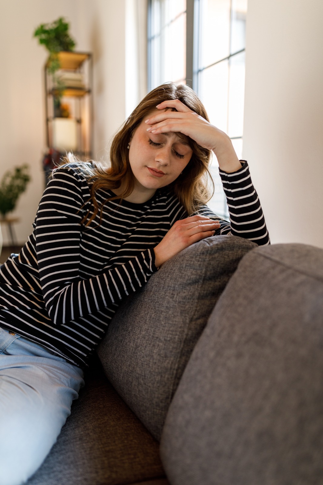 A young woman sitting on a couch with her head in her hand, looking thoughtful and worried